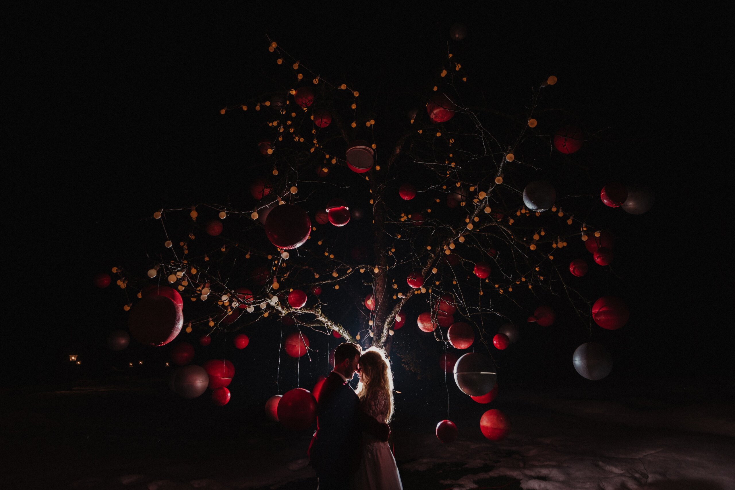 A couple embracing under a tree hung with glowing lights and red baubles at night